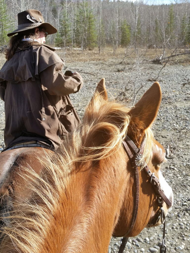 Rider in a long coat beside a chestnut horse on a gravel bar